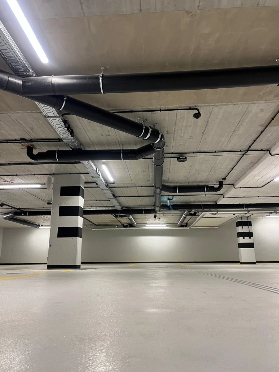 Nowoczesna podziemna hala garażowa z widocznymi czarnymi rurami instalacji sanitarnych zawieszonymi pod stropem